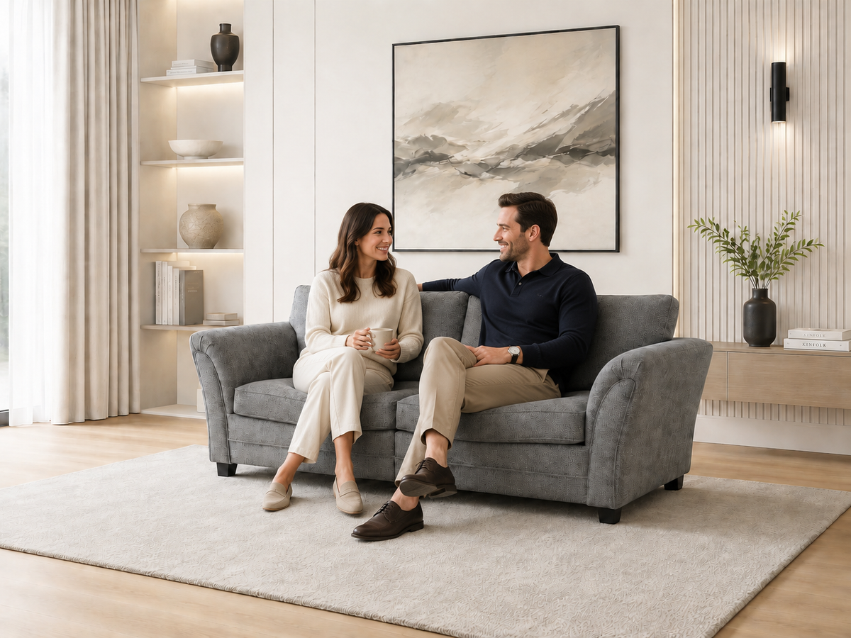 A man and a woman sit on a gray upholstered loveseat sofa